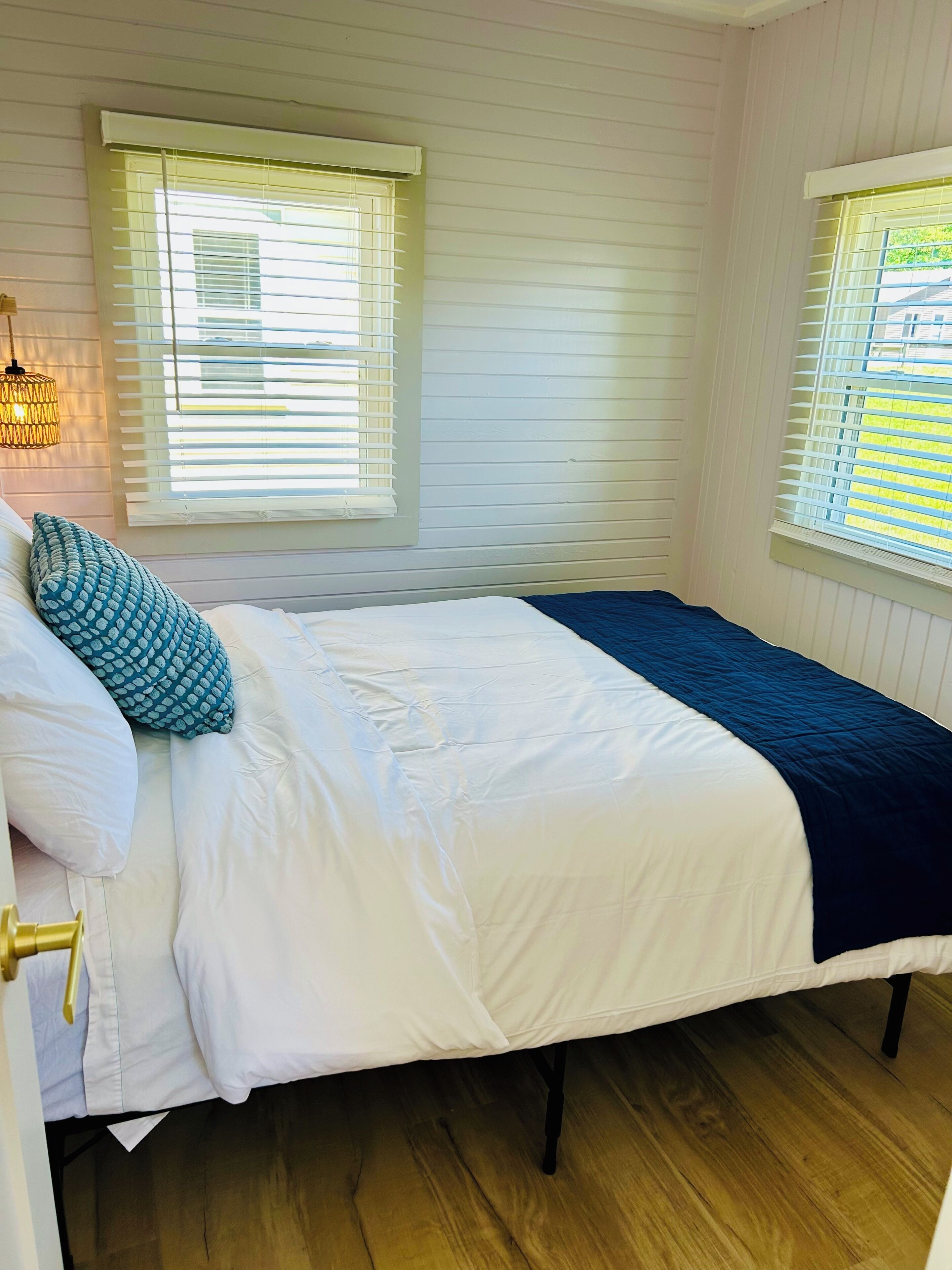 Une chambre à coucher avec un lit, deux fenêtres équipées de stores, un luminaire suspendu et un plancher en bois.