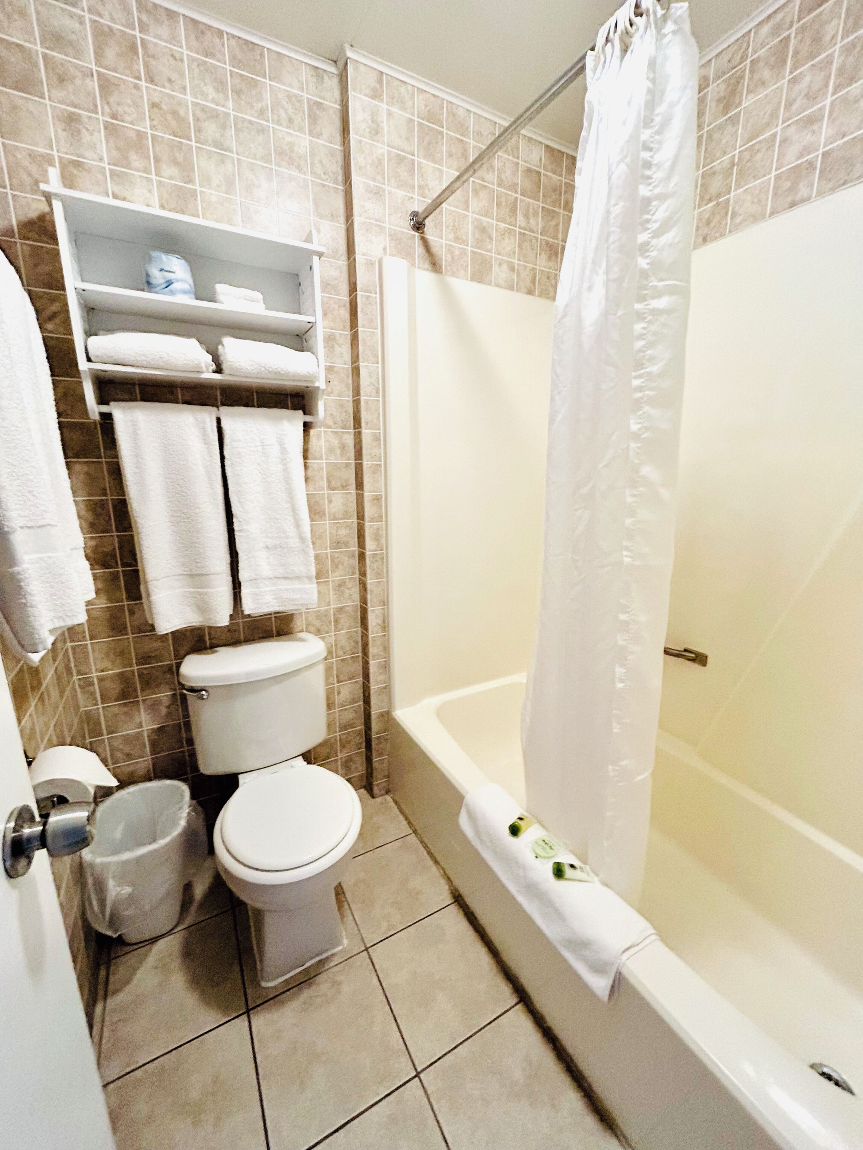 A bathroom with a toilet, a bathtub, and a shower curtain. There are towels hanging on a rack and a trash bin.