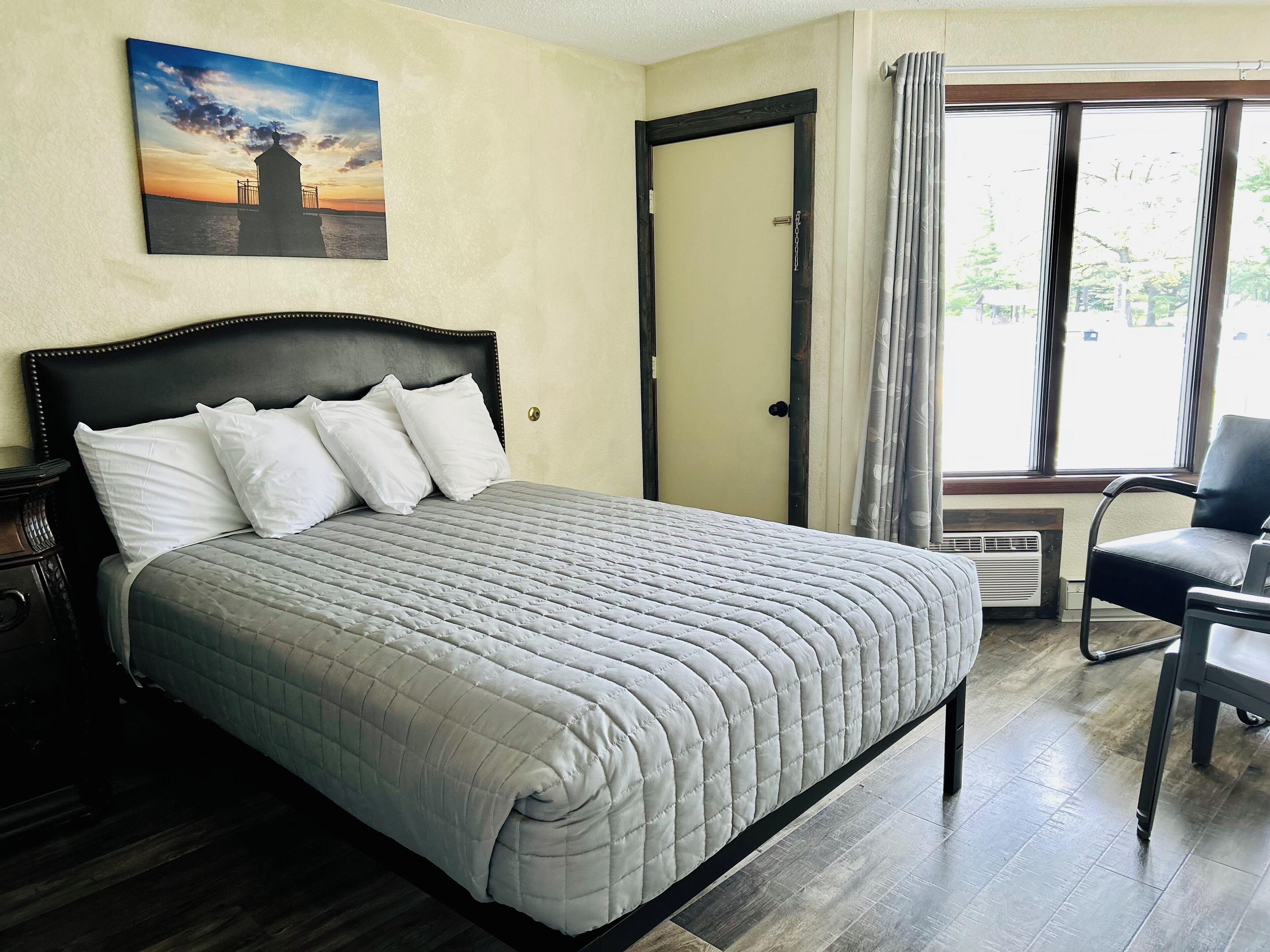 A neatly made bed with a grey quilt, a dark headboard, and white pillows. A painting of a sunset over a pier is on the wall.