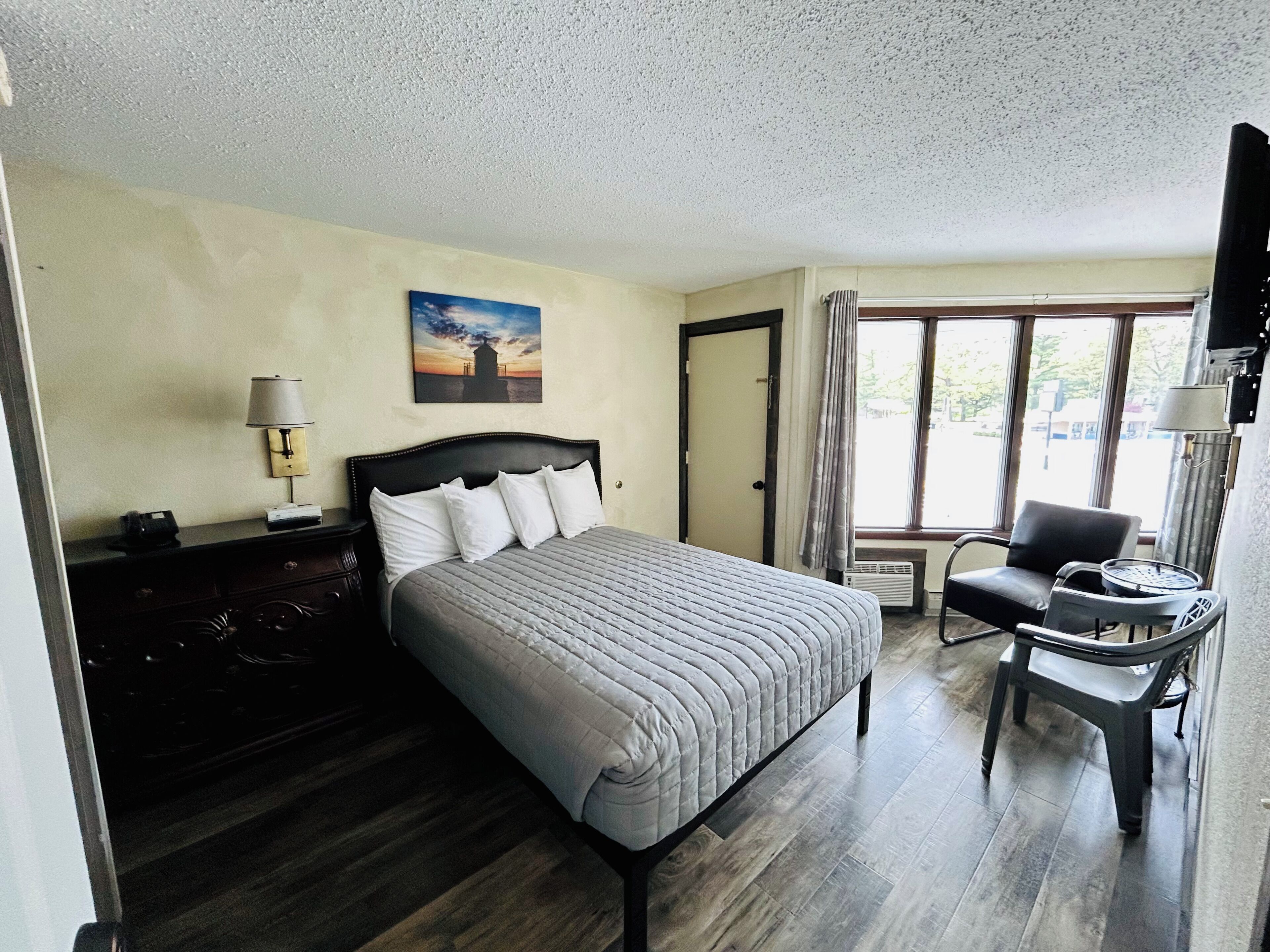 Habitación de hotel con cama, escritorio, silla, lámpara, un cuadro y ventana.
