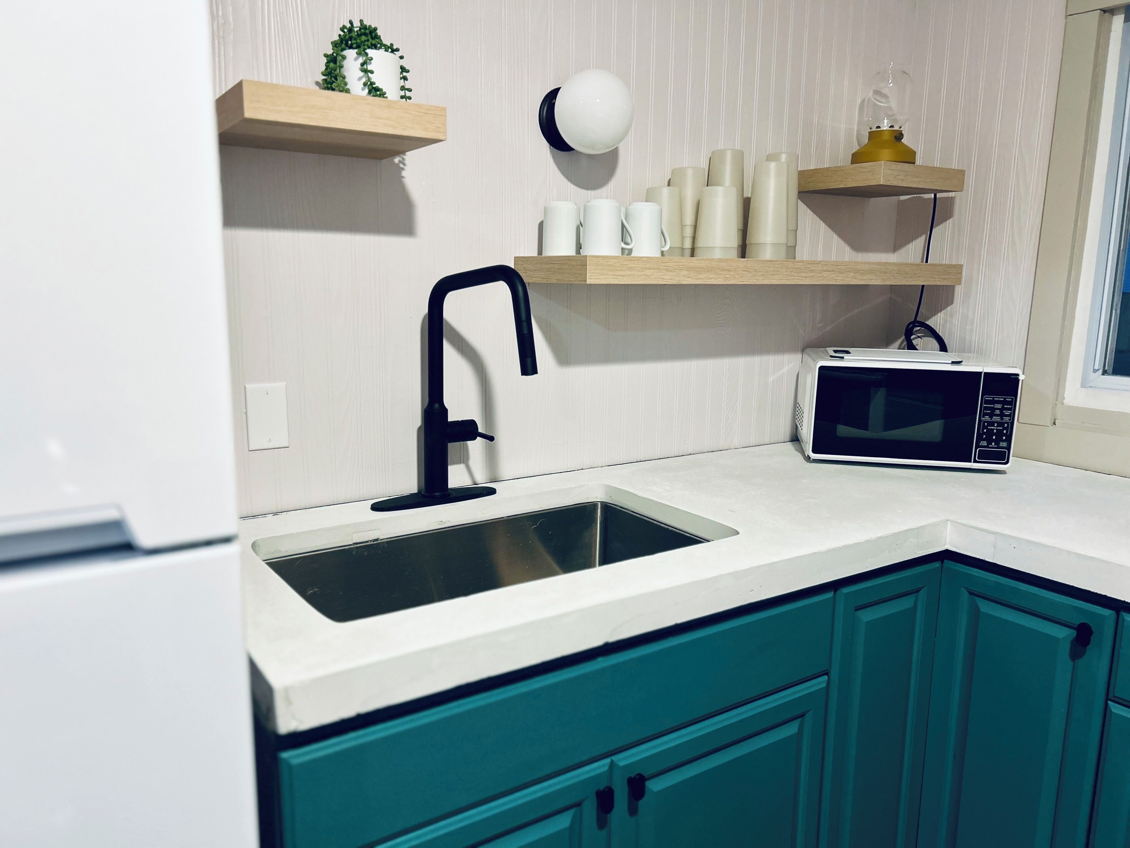 A kitchen with teal cabinets, a black faucet, a microwave, and shelves with cups and a plant.