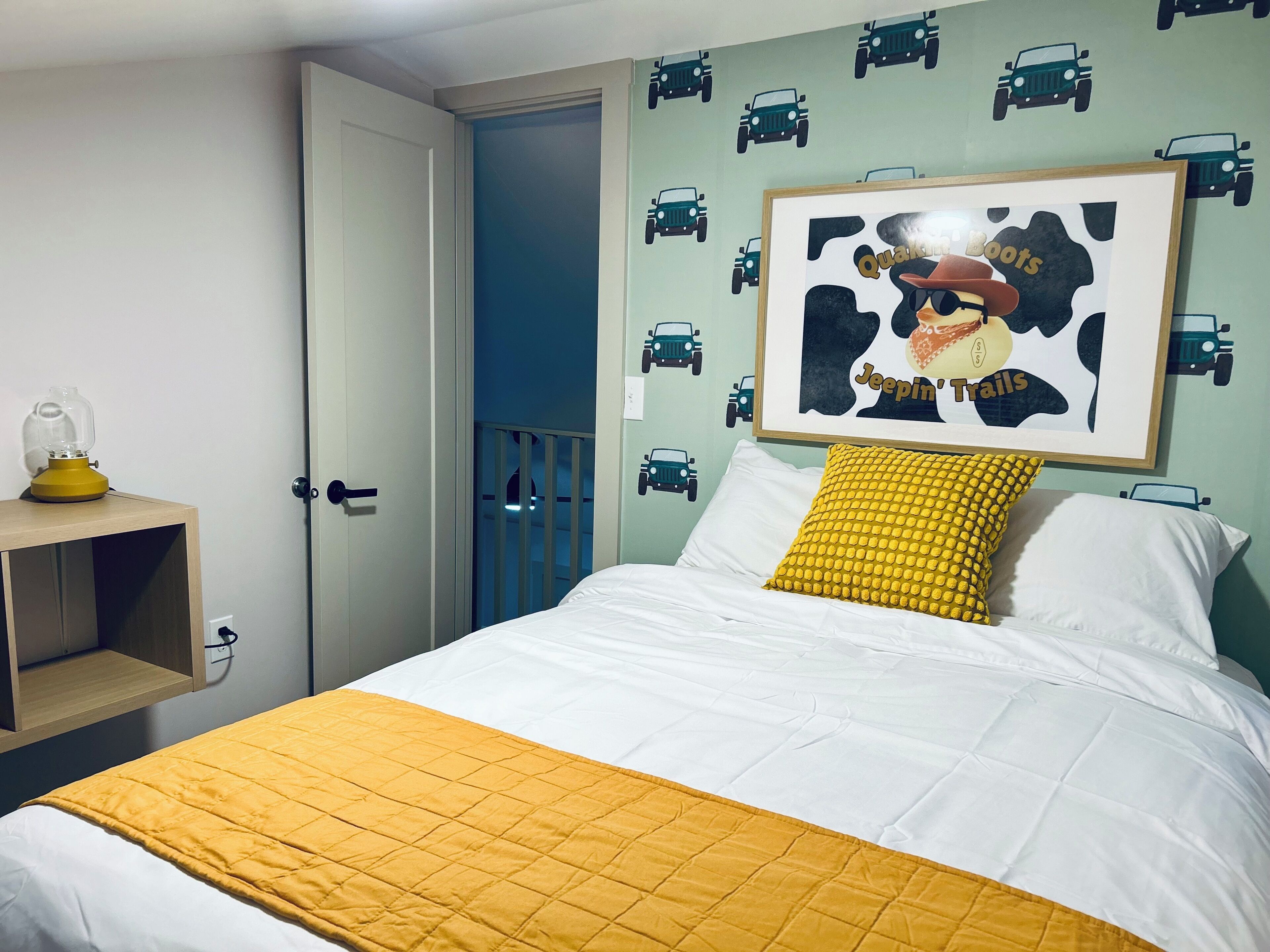 Une chambre à coucher avec un lit, un oreiller jaune, un tableau encadré accroché au mur et une porte dotée d’une rambarde bleue.