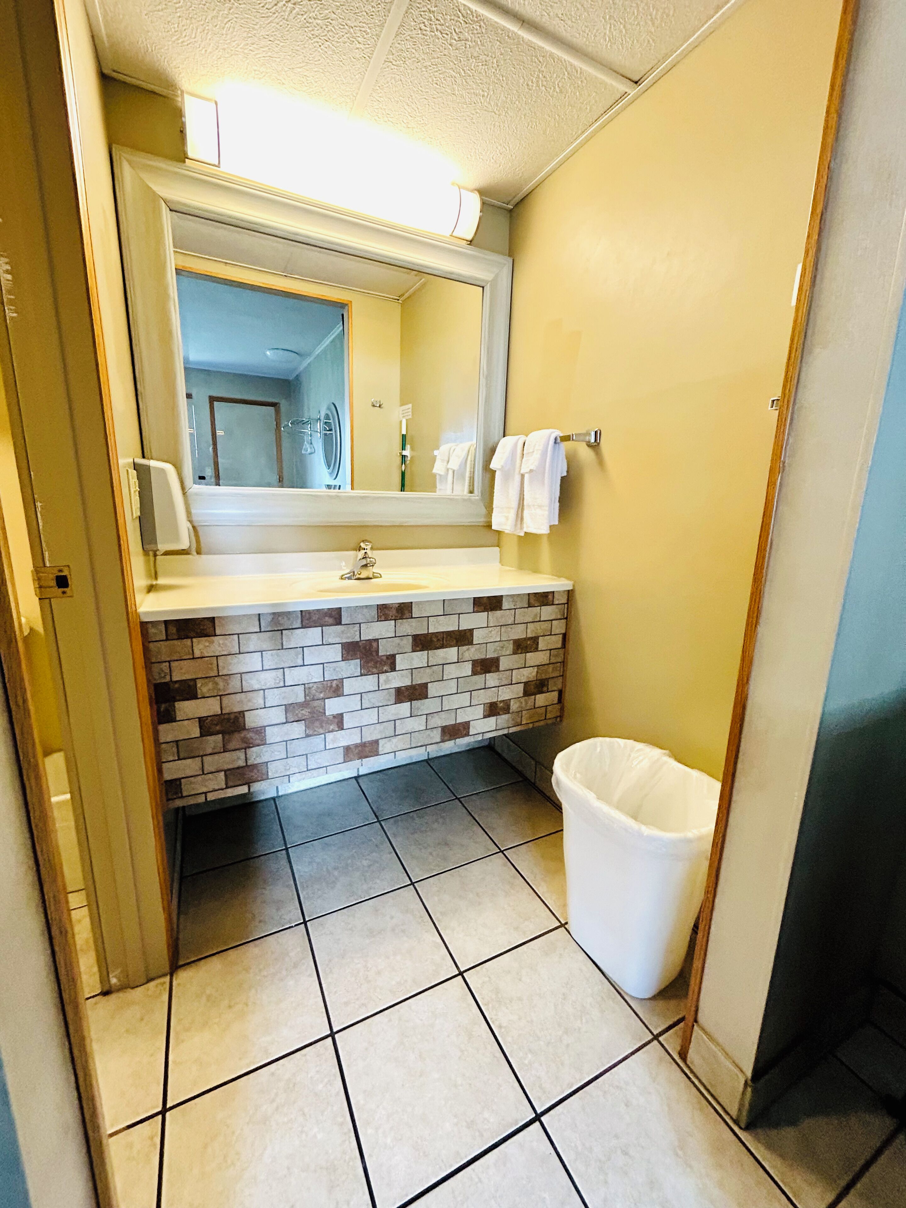 A bathroom with a tiled floor, a sink with a mirror, and a white trash bin.