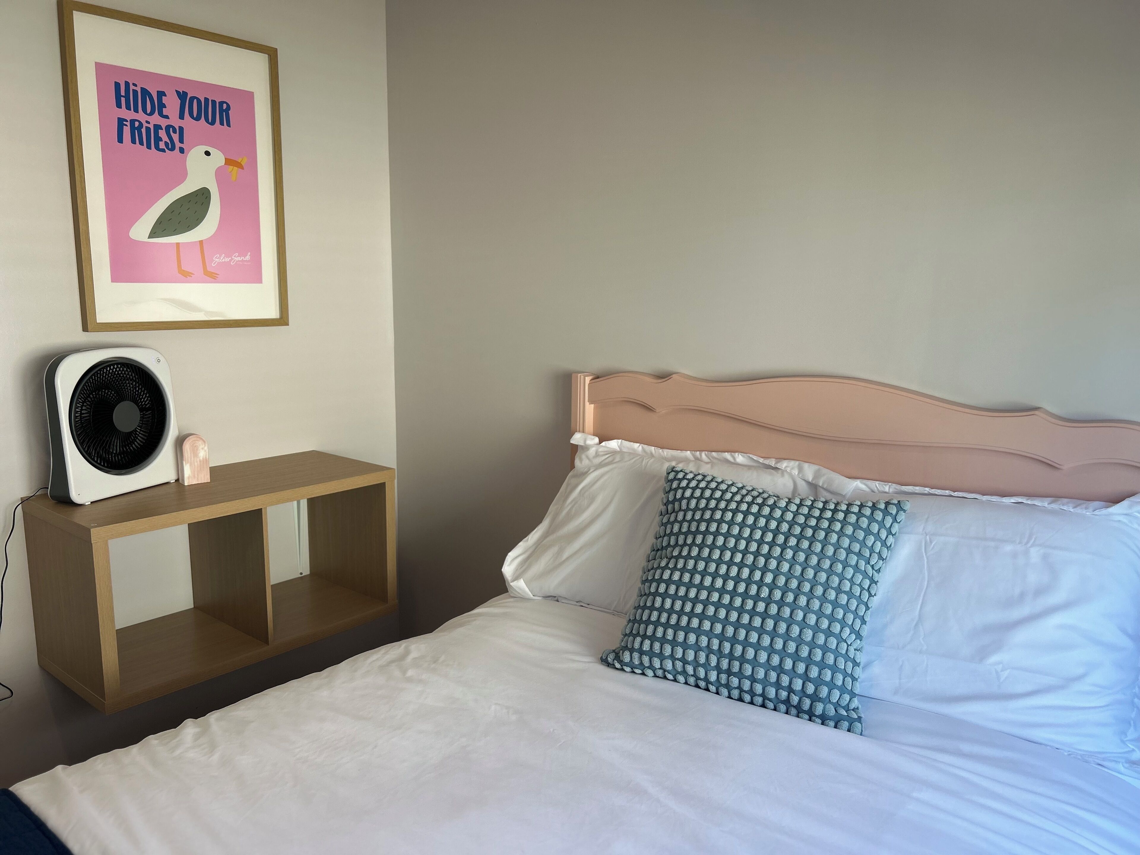 Een slaapkamer met een bed, kussens, een nachtkastje met een ventilator en een lijst met een poster aan de muur.