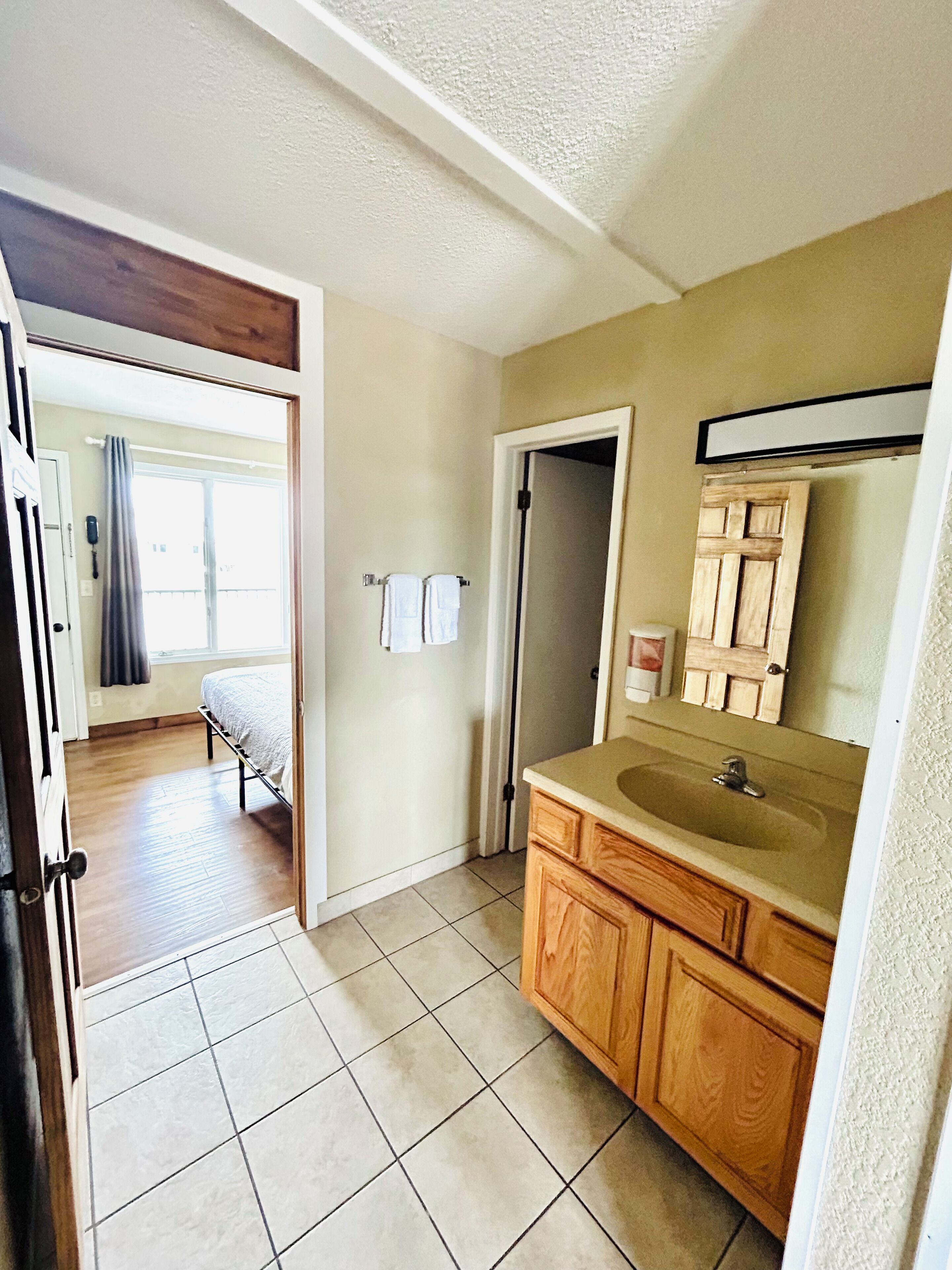 A bathroom with a wooden vanity, a sink, and a mirror. There is a door leading to a bedroom with a bed and a window.