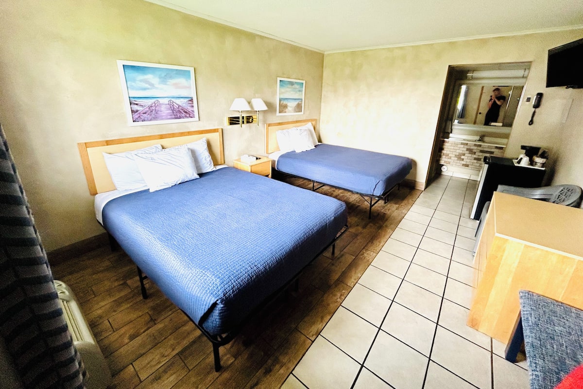 A hotel room with two beds, a wooden floor, a desk, and a bathroom visible through an open door.