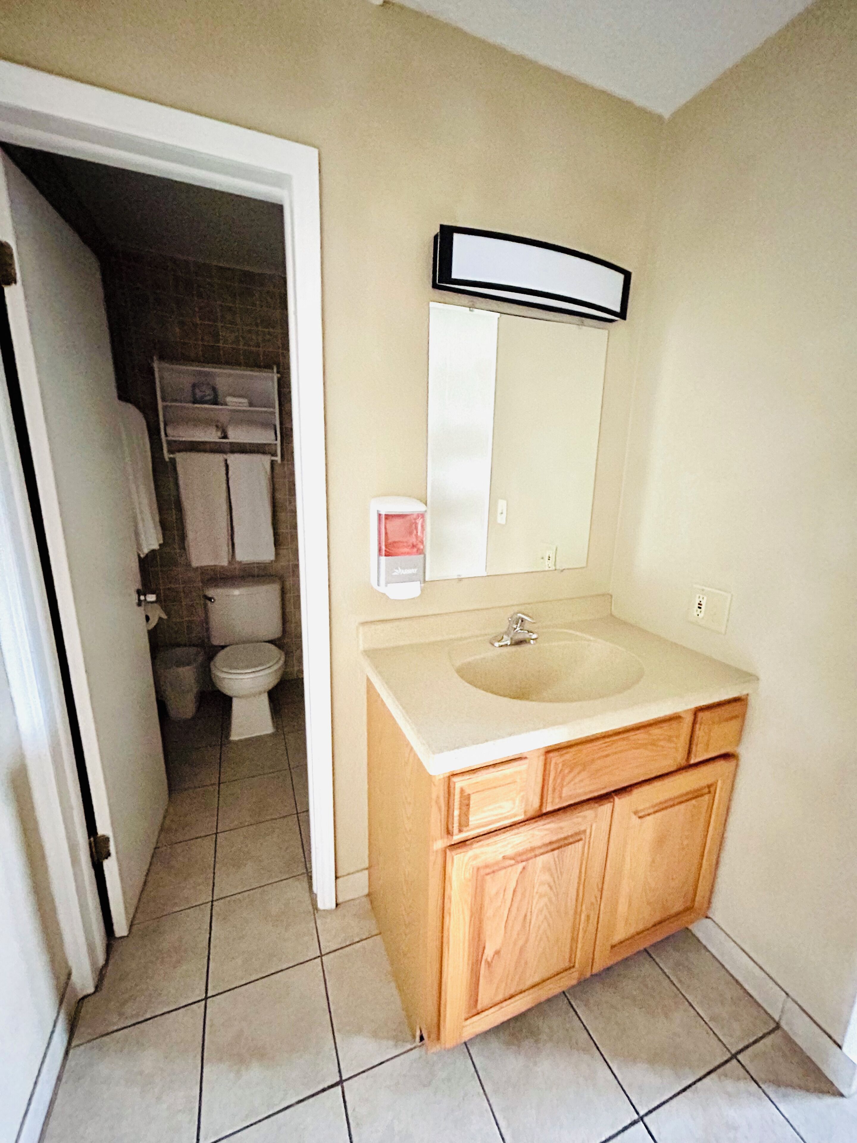 Baño con tocador de madera, lavamanos y espejo. Se ve un inodoro en la habitación contigua.