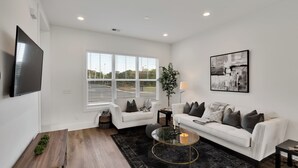 A modern living room with a white sofa, a glass coffee table, and a flat-screen TV mounted on the wall. - Stylish Two Story Close to South End and Uptown (Charlotte)