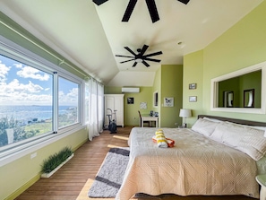A bedroom with a large bed, a ceiling fan, a view of the city, and a window.