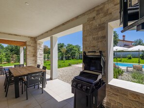 Um pátio externo com parede de pedra, churrasqueira, mesa de madeira com cadeiras e vista para uma piscina e vegetação.