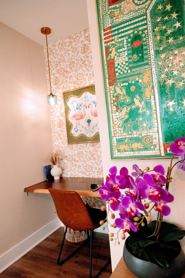 Una habitación con un escritorio de madera, una silla, una lámpara colgante, un papel tapiz con motivos y una planta en maceta con flores moradas.