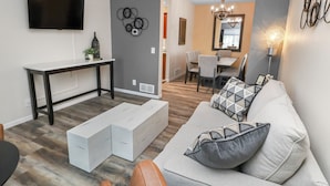 A modern living room with a sofa, a coffee table, and a TV mounted on the wall. There is a dining area with a table and chairs in the background.