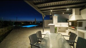 Une terrasse extérieure avec une table et des chaises en verre, un barbecue intégré et une vue sur une piscine au crépuscule.
