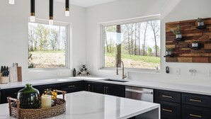 A modern kitchen with a large island, black cabinets, white countertops, and pendant lights. There are two windows providing views of a wooded area. - The Panorama Overlooking Center Hill Lake (Sparta)
