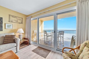 A room with a sofa, a small table, a lamp, and a view of the balcony with chairs and a table.