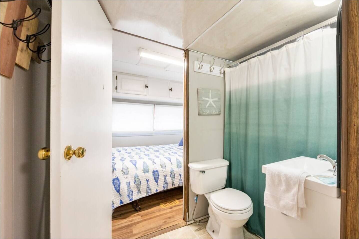 A compact, well-lit bathroom with a toilet, sink, and shower area. A bed with a patterned bedspread is visible in the adjacent room.