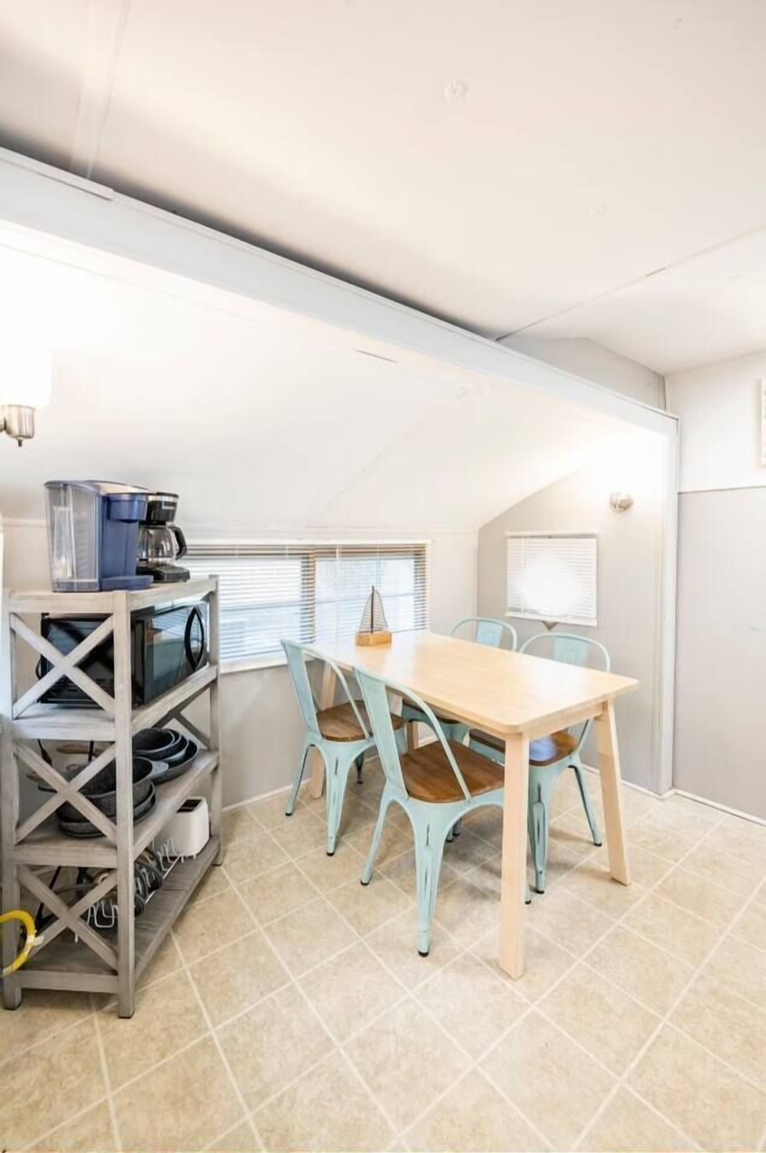 Une petite pièce avec une table en bois et quatre chaises, une machine à café posée sur une étagère et une fenêtre avec des stores.