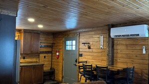 A cozy cabin interior with a wooden ceiling, a dining area with a table and chairs, a kitchen with wooden cabinets, and a leather sofa.