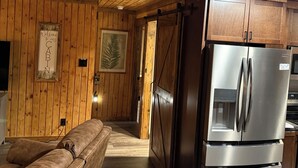 A cabin interior with wooden walls, a brown leather sofa, a stainless steel refrigerator, and a wooden door.