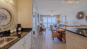 A modern kitchen with a large clock, a dining area with wicker furniture, and a view to the outside. - Calypso 404E, VIP Parking Spot, Beach Chairs! (Panama City Beach)