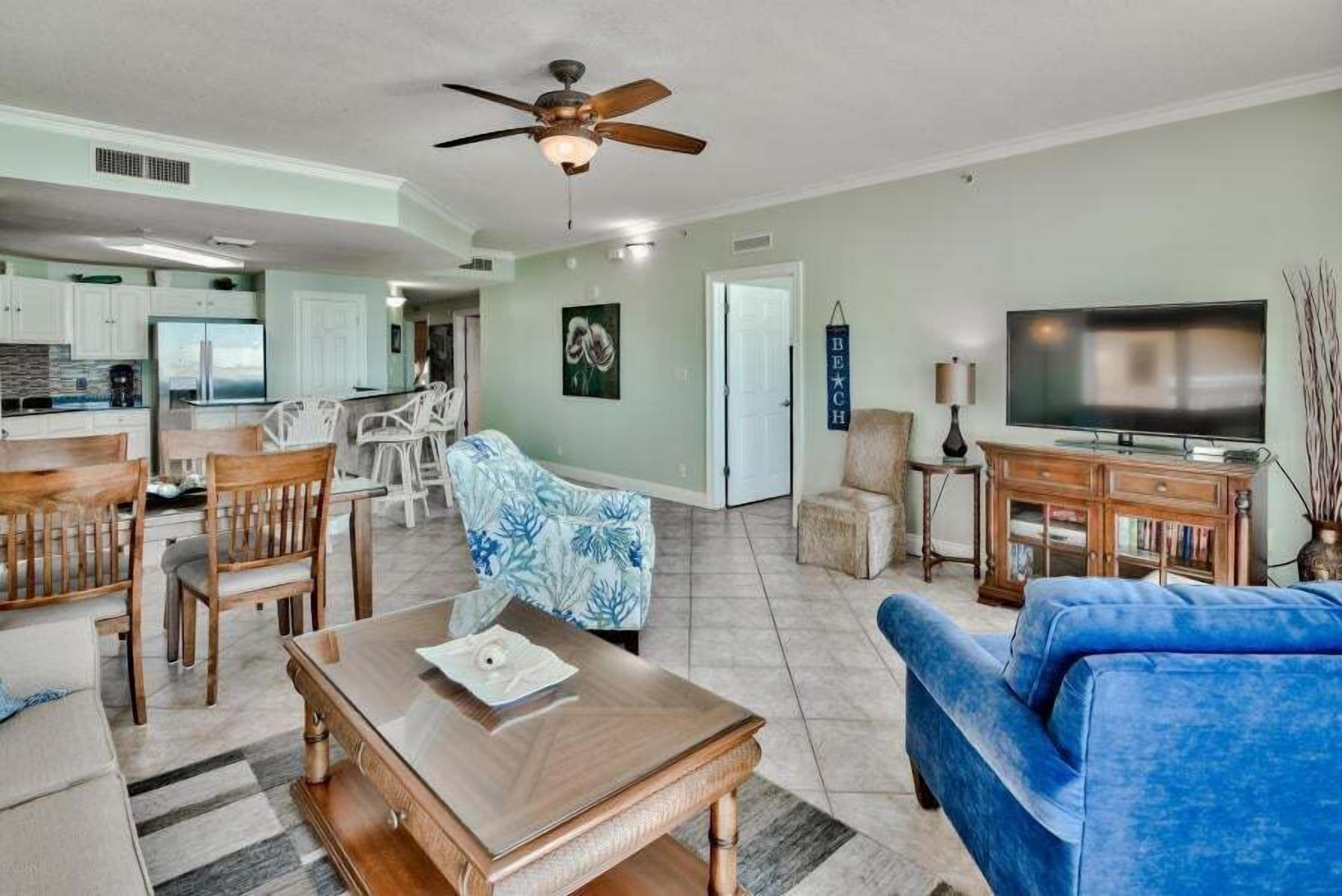 A spacious living area with a dining table, chairs, a blue sofa, a television, and a ceiling fan.