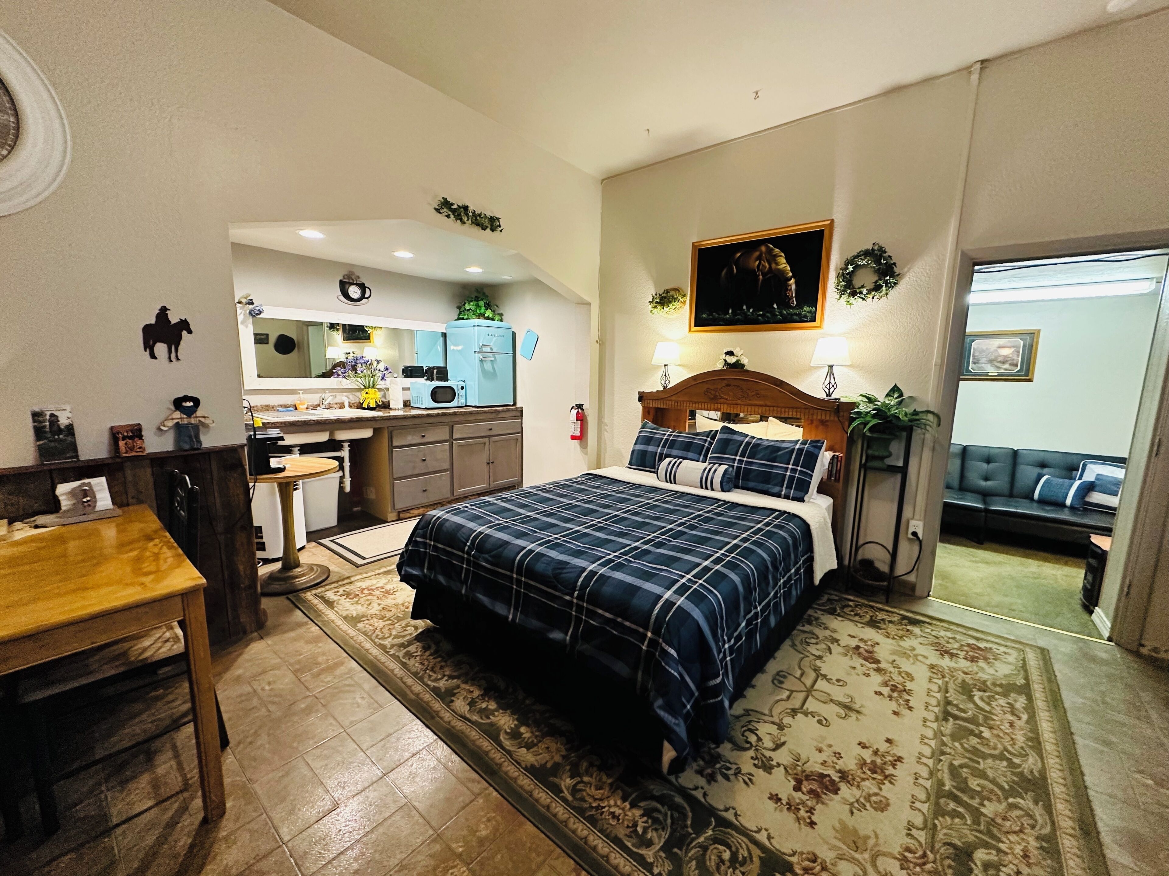 A bedroom with a bed, a desk, a chair, a sofa, and a kitchen area in the background.