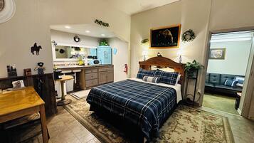 A bedroom with a bed, a desk, a chair, a sofa, and a kitchen area in the background.