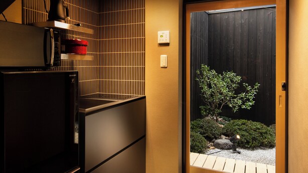 A modern kitchen with a built-in refrigerator, a microwave, and a coffee machine. There is a sliding door leading to a small garden with a bonsai tree. - Kanade Kiyomizu Gojo (Kyoto)