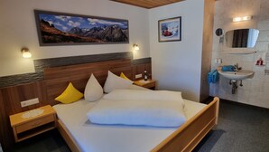A bedroom with a wooden headboard, a large bed with white bedding, two bedside tables with lamps, a wall-mounted painting, and a bathroom with a sink and mirror. - Vacation home Vesul by Interhome (Kappl)