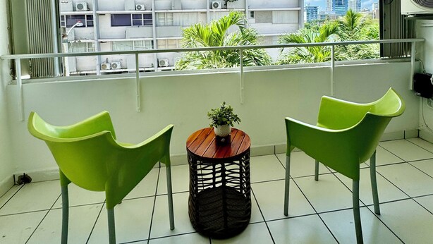 A balcony with two green chairs, a small wooden table with a plant, and a view of a building with multiple air conditioning units. - Condado Lagoon View @ Ritz Full Power Generator (San Juan)