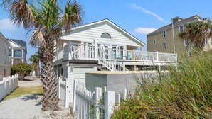Et hus ved stranden med balkon, trappe op til øvre etage og en sti af sand.