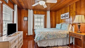 A bedroom with wooden walls, a bed with a floral bedspread, a TV on a dresser, and a ceiling fan. - Escape to Channel Charm: Relaxing Beach Getaway in North Myrtle's Cherry Grove with Scenic Views (North Myrtle Beach)