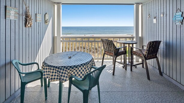 A balcony with a view of the ocean, featuring a round table and chairs, wooden walls, and decorative items.