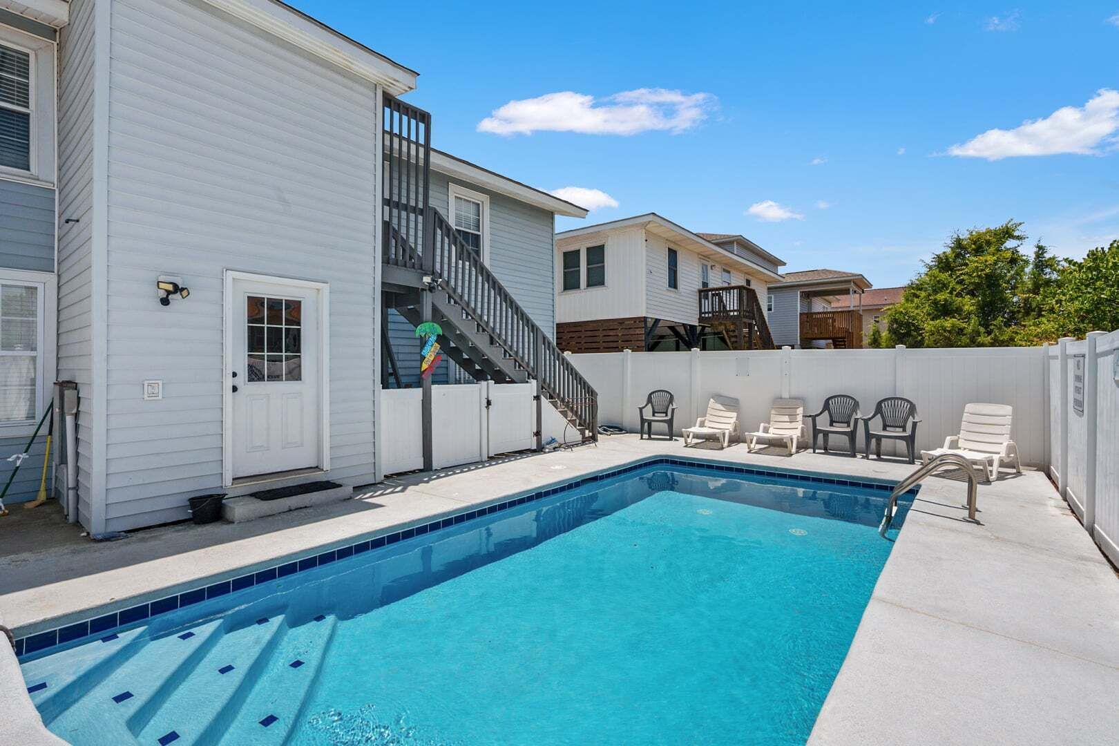 A swimming pool with a staircase leading to a deck area, surrounded by lounge chairs and a white building.