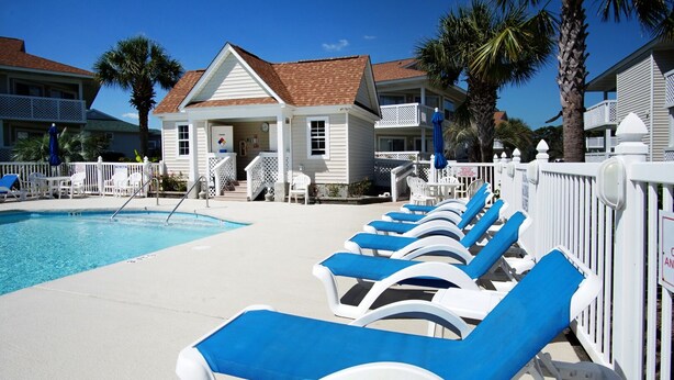 A pool area with lounge chairs and a small building with a white fence.