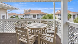 A covered outdoor patio with a round table and wooden chairs, white lattice railings, and a view of residential buildings in the background.