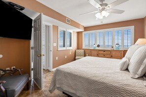 Un dormitorio con una cama grande, ventilador de techo, ventana con persianas, cómoda y una silla.