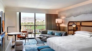 A hotel room with a large bed, a sofa, a dining table, and chairs. There is a view of mountains and greenery through the large windows. - Marriott Desert Spring Villas- available march 2-9 only (palm desert)