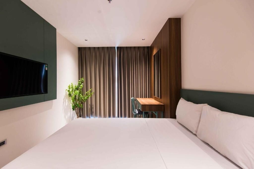 A modern hotel room with a large bed, a flat-screen TV mounted on the wall, a small table with a chair, and a large window with curtains.