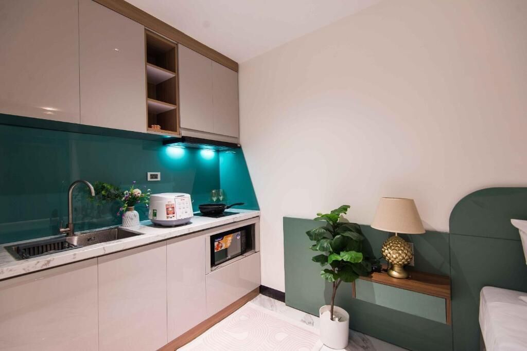 Een kleine keuken met een spoelbak, magnetron en een klein aanrecht. Er staat een groene plant en een lamp op een nachtkastje.