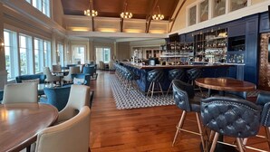 A spacious bar area with high ceilings and large windows, featuring a bar counter, stools, and a seating area with tables and chairs. - High Tide Hideaway by Sandpiper | Near Beach (Seabrook Island)