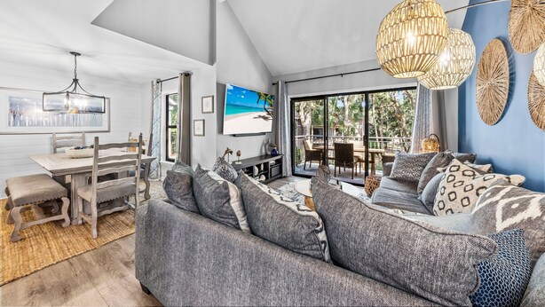 A modern living room with a large sofa, a dining table, and a TV. - High Tide Hideaway by Sandpiper | Near Beach (Seabrook Island)