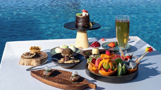 A dining table with a variety of dishes and a glass of drink, overlooking a city skyline and a body of water.