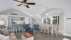 A spacious living area with a high ceiling, a dining table with chairs, a kitchen with light blue cabinets, and a large window offering a view of trees. - Cape Cod Magic at Starfish Cottage (South Yarmouth)