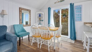 Un comedor con una mesa de madera y sillas blancas, un sillón azul y un armario blanco con estantes.