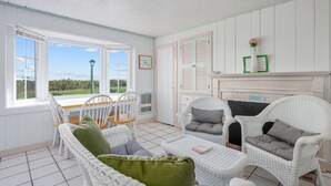 A bright, white room with a large window, wicker furniture, and a fireplace.