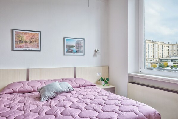 A bedroom with a pink quilted bedspread, a bedside table with a plant, and two framed pictures on the wall.