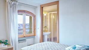 Une chambre à coucher avec un lit, une fenêtre avec des rideaux, un radiateur, une table de chevet avec une plante, et une salle de bain visible à travers une porte ouverte.