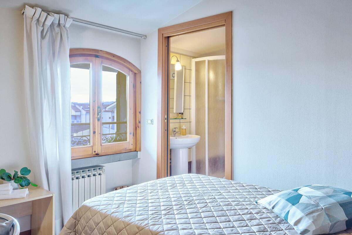 Quarto com cama, janela com cortinas, radiador, mesa de cabeceira com planta e banheiro visível por uma porta aberta.