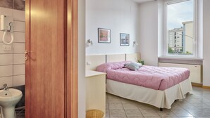 Une chambre à coucher avec un lit, une table de chevet, une fenêtre et une vue sur les bâtiments à l’extérieur.